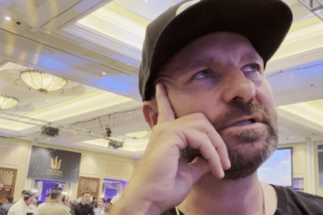 Mies mustassa lippiksessä nojaa käteensä ja katsoo yläviistoon. Takana näkyy muita ihmisiä istumassa pöytien äärellä. Oikealla seisoo kaksi miestä.