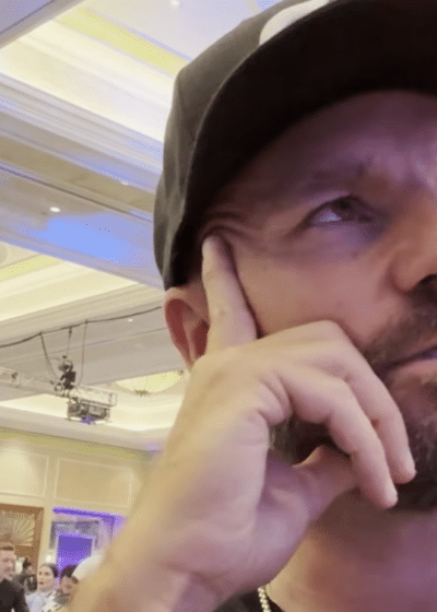 Mies mustassa lippiksessä nojaa käteensä ja katsoo yläviistoon. Takana näkyy muita ihmisiä istumassa pöytien äärellä. Oikealla seisoo kaksi miestä.