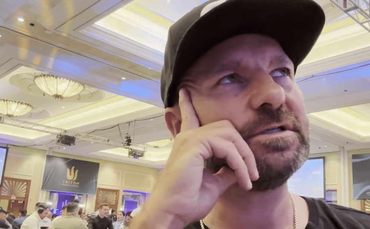 Mies mustassa lippiksessä nojaa käteensä ja katsoo yläviistoon. Takana näkyy muita ihmisiä istumassa pöytien äärellä. Oikealla seisoo kaksi miestä.