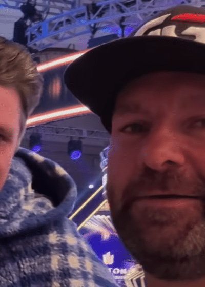 Kuvassa on kaksi miestä, toisella on musta lippalakki päässään. Molemmat hymyilevät ja katsovat kameraan. Taustalla näkyy sinistä ja mustaa.