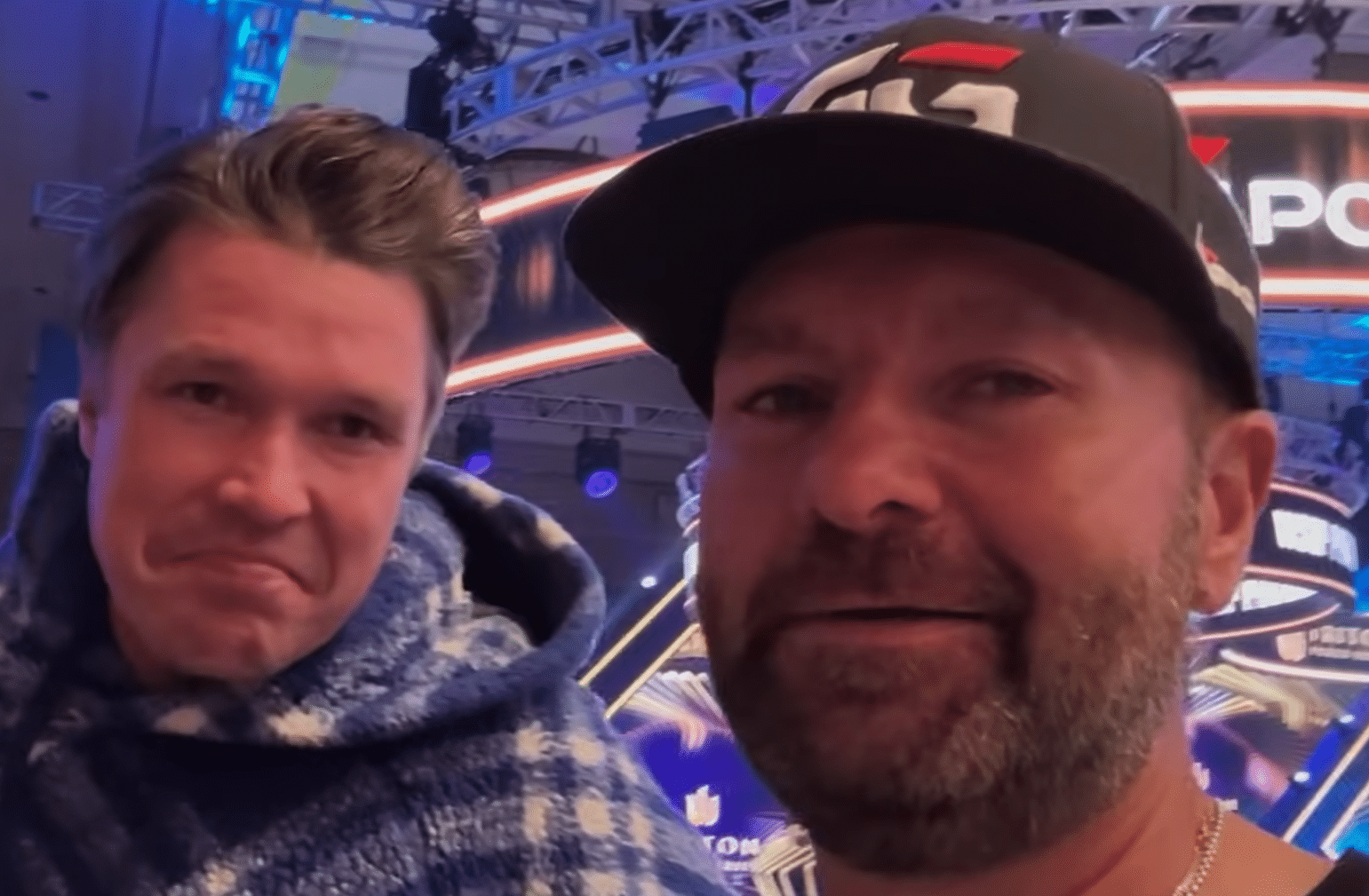 Kuvassa on kaksi miestä, toisella on musta lippalakki päässään. Molemmat hymyilevät ja katsovat kameraan. Taustalla näkyy sinistä ja mustaa.