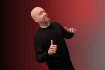 Kalju mies seisoo kuvassa. Hänellä on päällään tumma paita. Miehellä on molempien käsien peukalot pystyssä. Tausta on punainen.