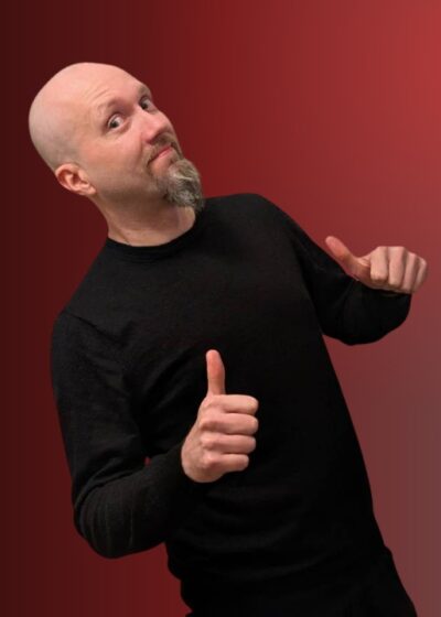 Kalju mies seisoo kuvassa. Hänellä on päällään tumma paita. Miehellä on molempien käsien peukalot pystyssä. Tausta on punainen.