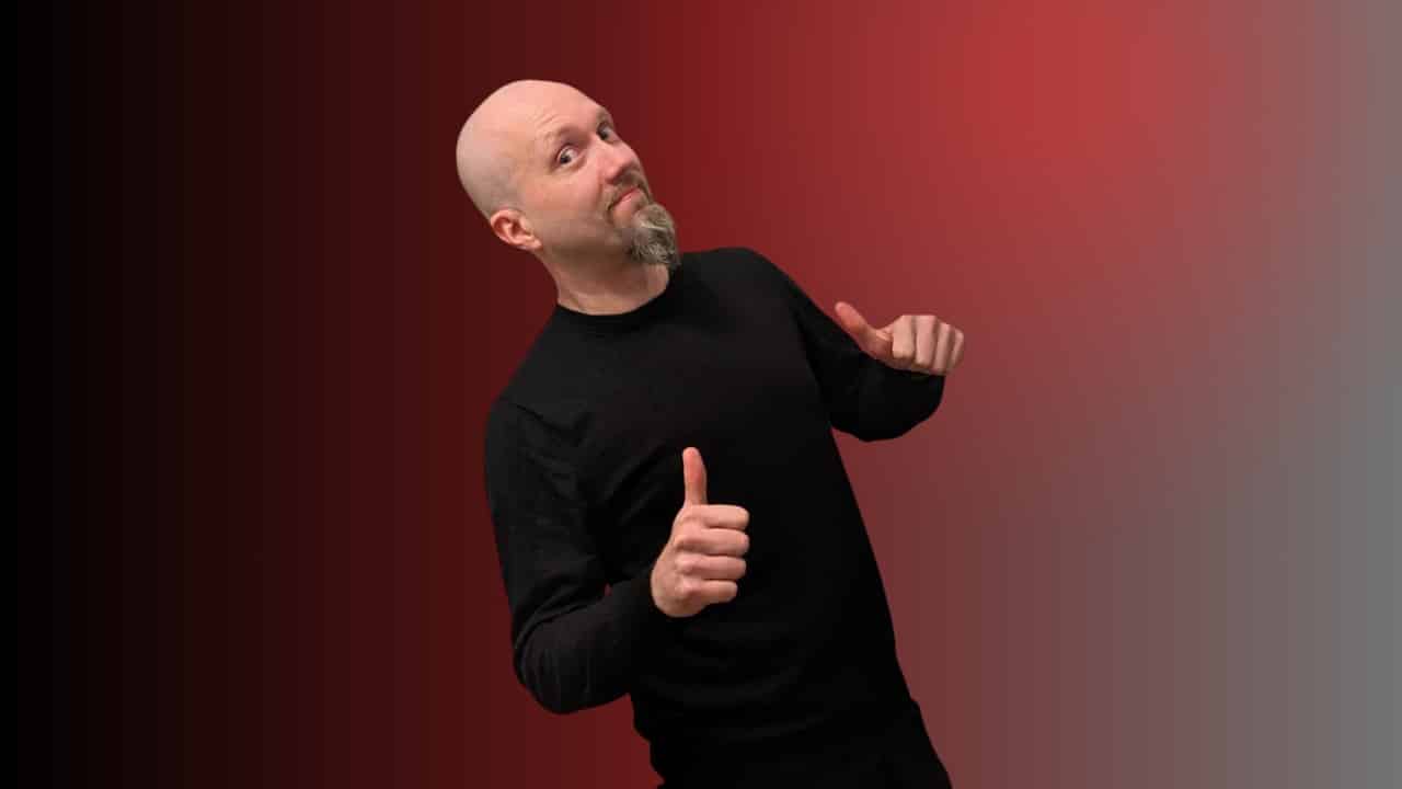 Kalju mies seisoo kuvassa. Hänellä on päällään tumma paita. Miehellä on molempien käsien peukalot pystyssä. Tausta on punainen.