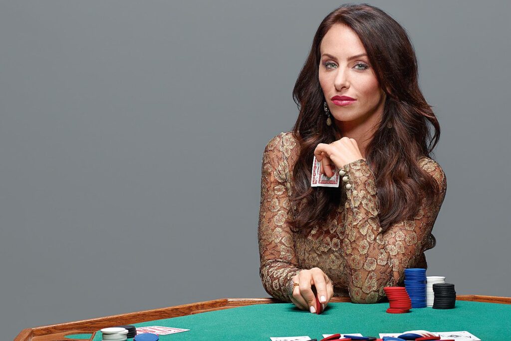 Nainen istuu pokeripöydässä. Hänellä on päällään kultainen mekko. Vihreällä pöydällä on erivärisiä pelimerkkejä.