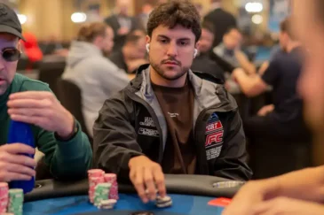 Kaksi miestä istuu pokeripöydässä. Pöydällä on erivärisiä pelimerkkejä. Toisella miehellä on aurinkolasit ja lippis päässä.