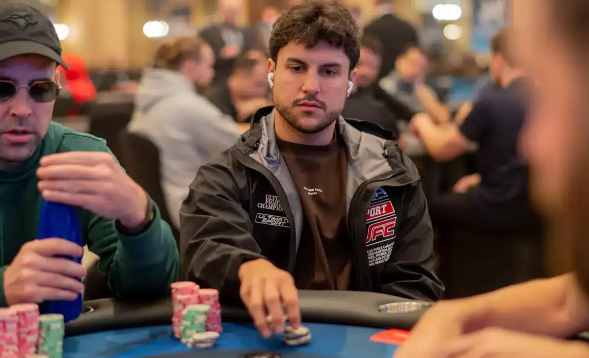 Kaksi miestä istuu pokeripöydässä. Pöydällä on erivärisiä pelimerkkejä. Toisella miehellä on aurinkolasit ja lippis päässä.