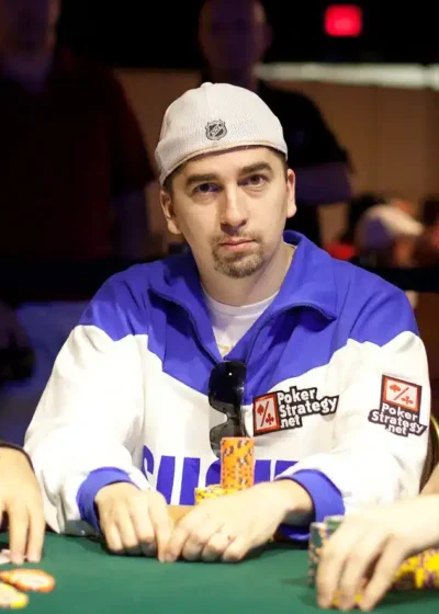 Kolme miestä istuu pokeripöydässä. Pöydässä on vihreä verka. Keskimmäisellä miehellä on lippis päässään ja sinivalkoinen paita.