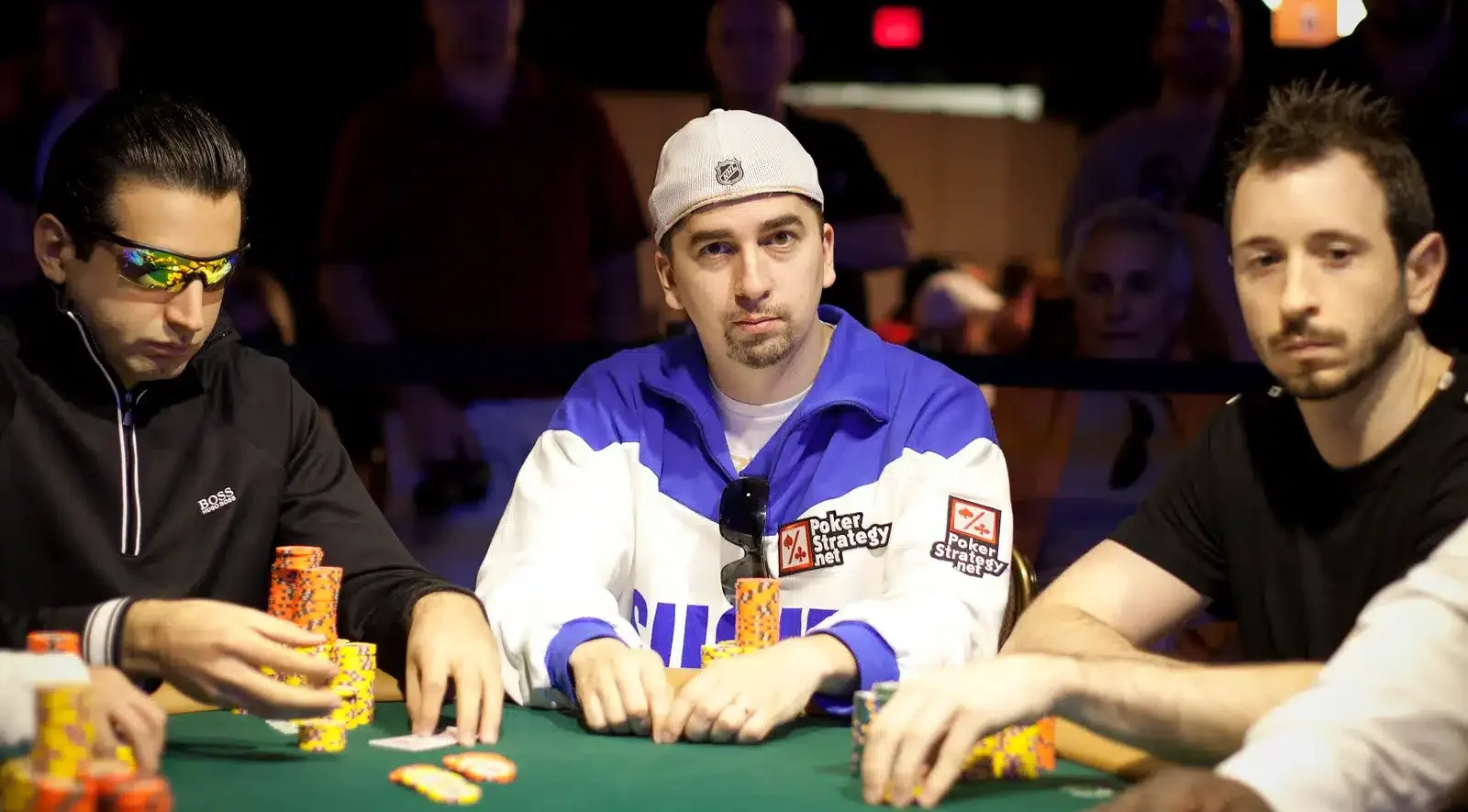 Kolme miestä istuu pokeripöydässä. Pöydässä on vihreä verka. Keskimmäisellä miehellä on lippis päässään ja sinivalkoinen paita.