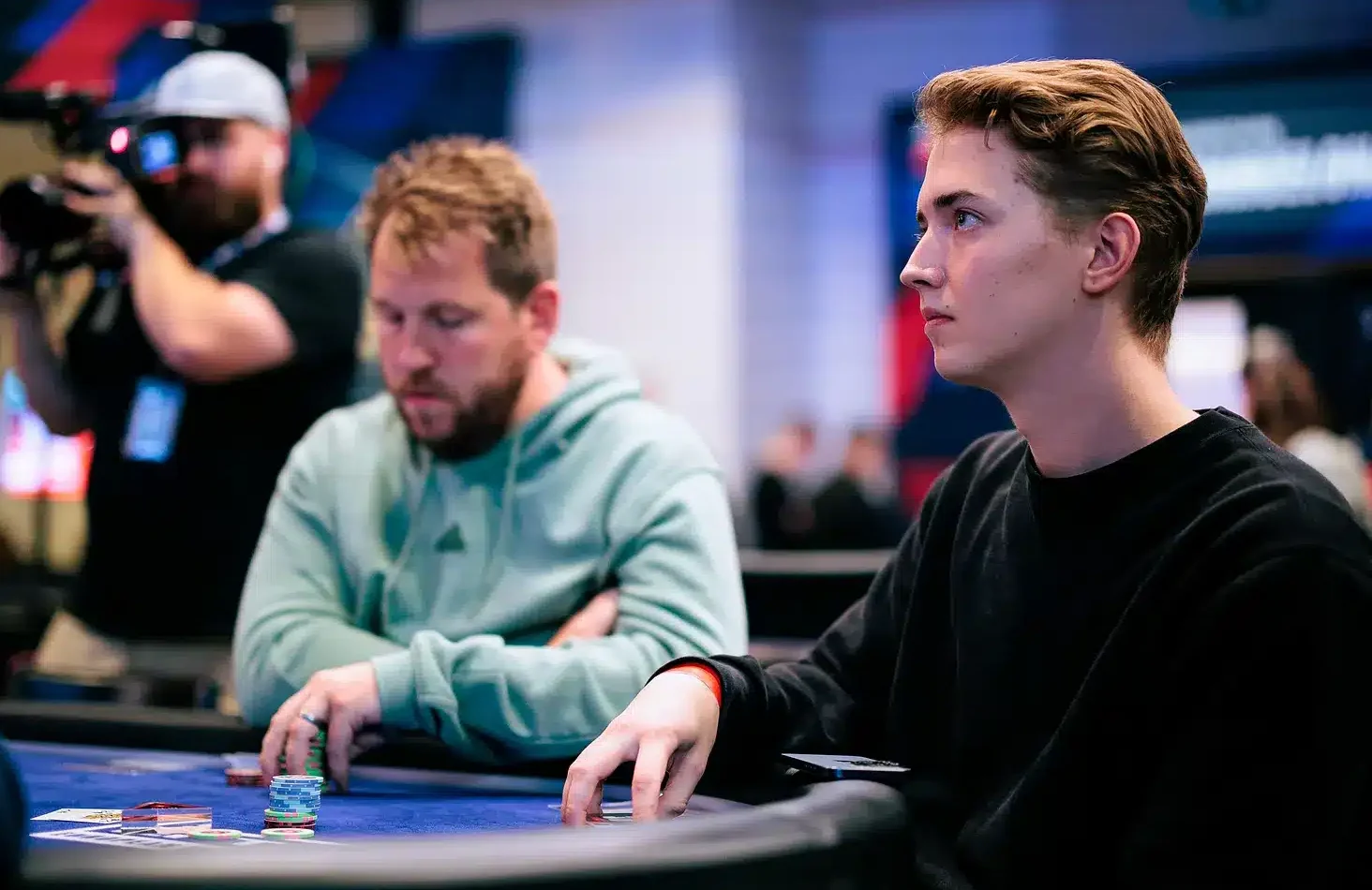 Luka Sorainen istumassa pelipöydässä vieressä vihreähupparinen mies ja takana kameramies