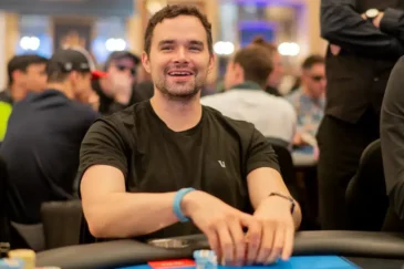 Alan Keating istuu pokeripöydässä ja hymyilee Hänellä on päällään musta t-paita. Hänellä on molemmat kädet pöydällä.