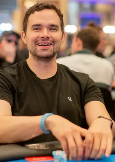 Alan Keating istuu pokeripöydässä ja hymyilee Hänellä on päällään musta t-paita. Hänellä on molemmat kädet pöydällä.