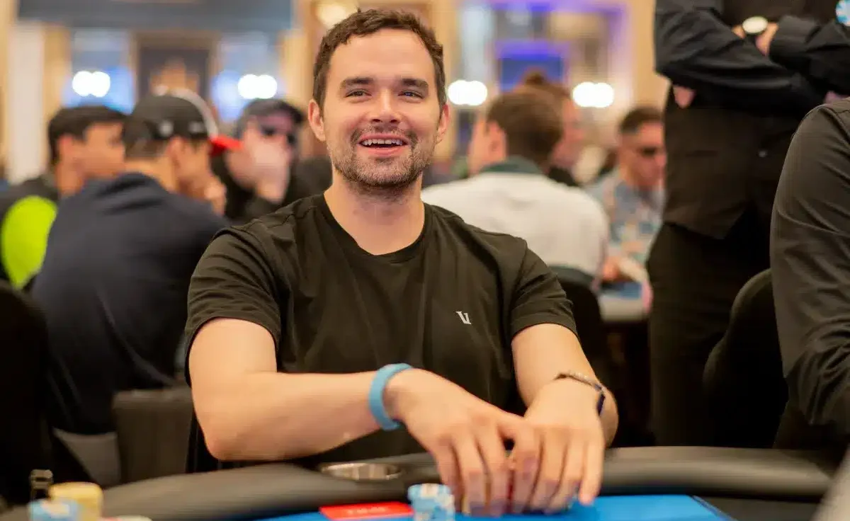 Alan Keating istuu pokeripöydässä ja hymyilee Hänellä on päällään musta t-paita. Hänellä on molemmat kädet pöydällä.