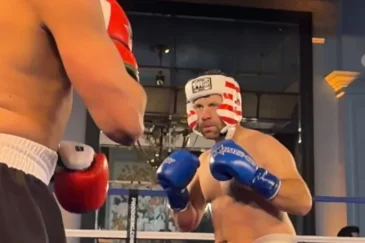 Mies nyrkkeilykehässä ilman paitaa. Kädessään siniset hanskat ja päässään punavalkoraitainen kypärä.