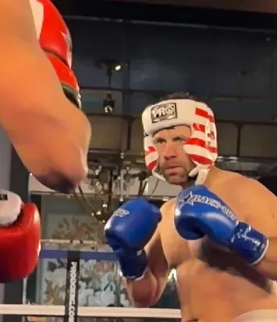 Mies nyrkkeilykehässä ilman paitaa. Kädessään siniset hanskat ja päässään punavalkoraitainen kypärä.