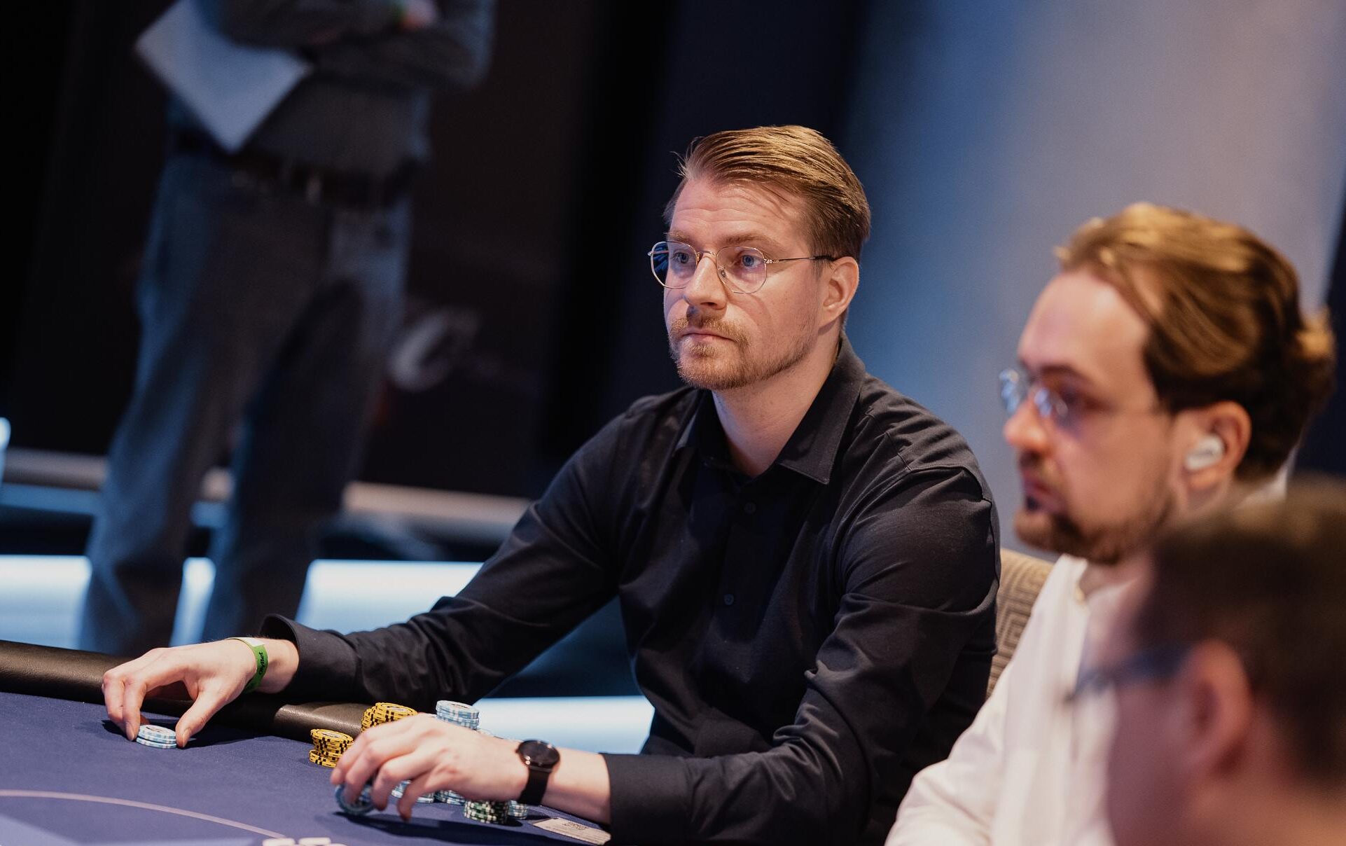 Mies tumman sinisessä kauluspaidassa istuu sinisen pokeripöydän äärellä