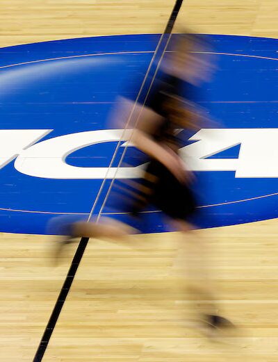 Kuvassa on koripallokentän parketti, jossa on sininen "NCAA"-logo. Logon ohi juoksee mies.