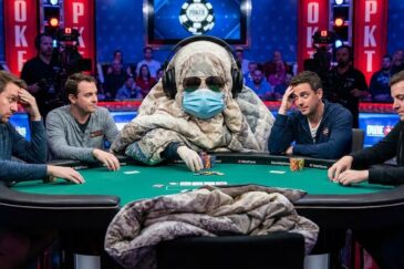 Kuvassa on pokeripöytä, jossa istuu keskellä peittoon kääriytynyt ihminen, jolla on aurinkolasit ja kasvomaski.