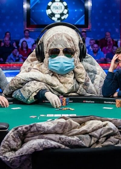 Kuvassa on pokeripöytä, jossa istuu keskellä peittoon kääriytynyt ihminen, jolla on aurinkolasit ja kasvomaski.