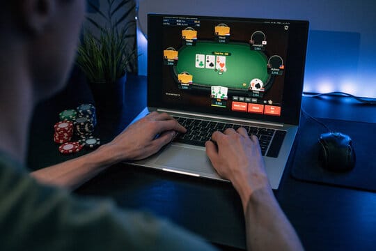 Mies pelaamassa nettipokeria tummssa huoneessa. Pöydällä on tietokone ja pelimerkkejä, sekä hiiri.
