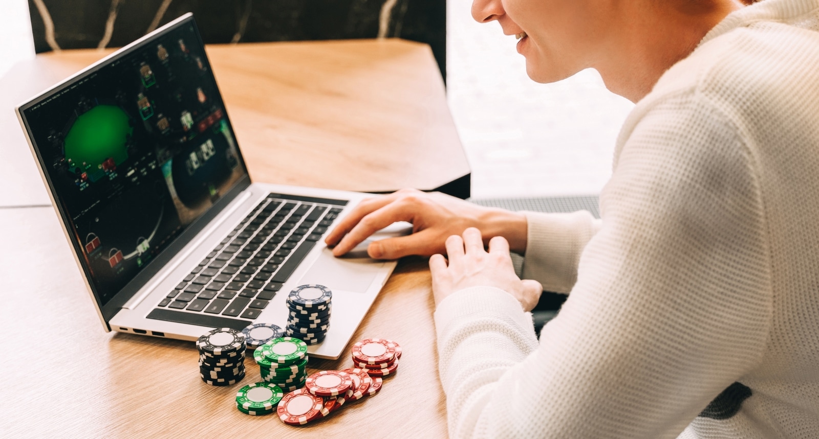 Kuvassa mies pelaa tietokoneella pokeria, vieressä hänellä on kasa pelimerkkejä.