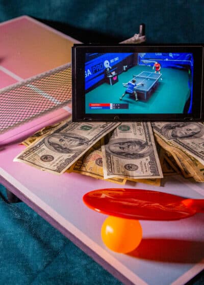 Kuvassa on pieni pingispöytä, jonka päällä on kaksi mailaa, pallo ja seteleitä, sekä tabletti.