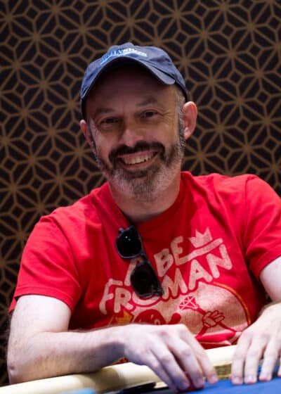 Mies sinisessä lippiksessä ja punaisessa t-paidassa hymyilee pokeripöydän äärellä.