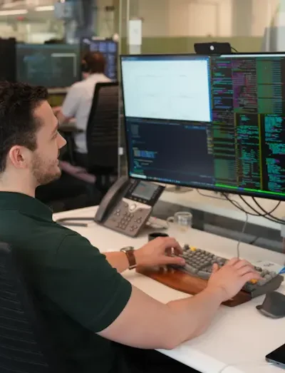 Kuvassa on mies monen tietokonenäytön edessä pöydän äärellä. Miehellä on musta t-paita