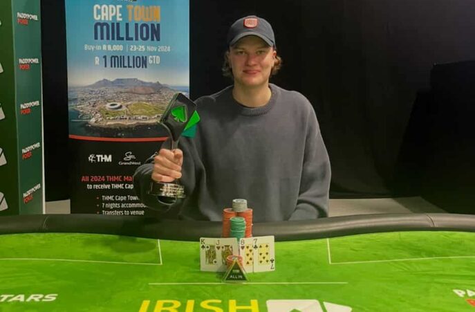 Toivo Rinne istuu pokeripöydässä. Hänellä on harmaa paita päällään ja päässään lippis. Kädessään hänellä on pokaali.