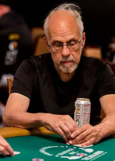 Harmaahiuksinen mies istuu pokeripöydässä. Hänen edessään pöydällä on vaalea tölkki. Pöydän pinta on vihreän värinen.