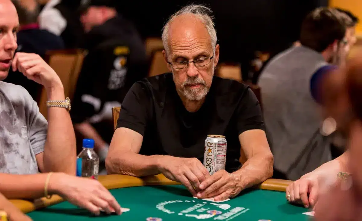 Harmaahiuksinen mies istuu pokeripöydässä. Hänen edessään pöydällä on vaalea tölkki. Pöydän pinta on vihreän värinen.