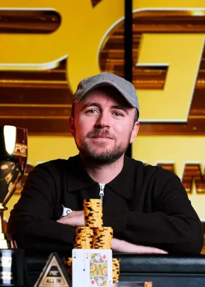 Patrick Leonard istuu pokeripöydässä. Hänellä on päällään musta paita ja lippis. Hänen edessään pöydällä on voittopokaali.