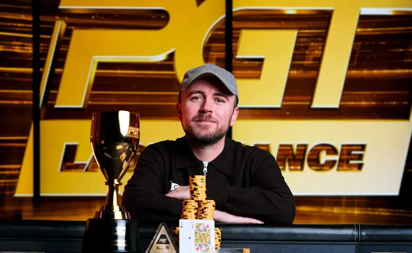 Patrick Leonard istuu pokeripöydässä. Hänellä on päällään musta paita ja lippis. Hänen edessään pöydällä on voittopokaali.