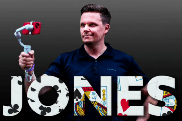 Miehellä on kädessään puhelin, joka on telineessä kiinni. Hänellä on tumman sininen paita, tausta on tumma