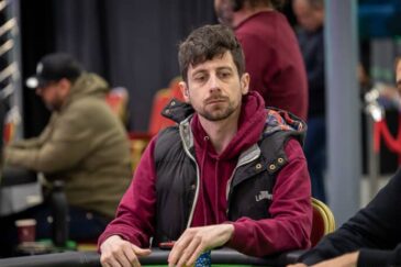 Mies istuu pöydän ääressä punaisessa hupparissa ja mustassa liivissä. Pöytä on vihreä, hänen vieressään istuu toinen mies.