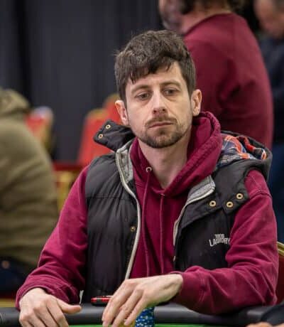 Mies istuu pöydän ääressä punaisessa hupparissa ja mustassa liivissä. Pöytä on vihreä, hänen vieressään istuu toinen mies.