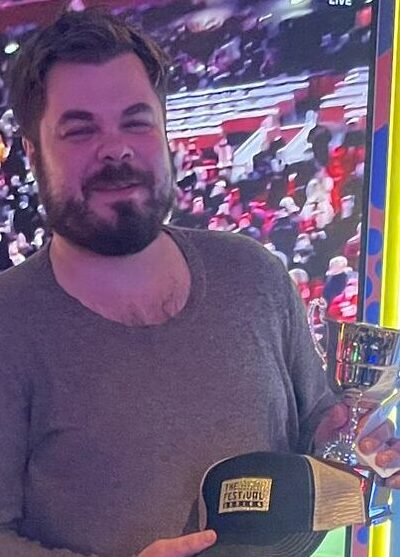 Mies seisoo ison televiison edessä. Hänellä Hänellä on tummat farkut ja ruskeat kengät. Kädessään mies pitää lippistä sekä pokaalia.