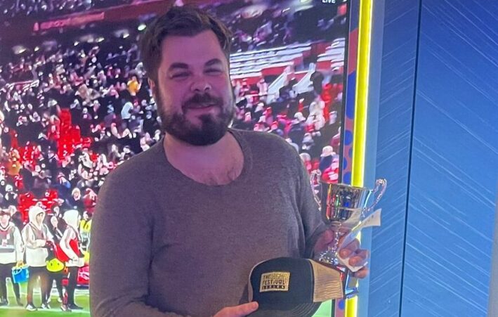 Mies seisoo ison televiison edessä. Hänellä Hänellä on tummat farkut ja ruskeat kengät. Kädessään mies pitää lippistä sekä pokaalia.