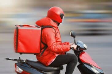 Kuvassa on ruokalähetti joka kulkee skootterilla päällään punainen takki ja punainen kypärä.