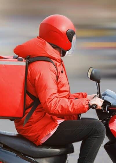Kuvassa on ruokalähetti joka kulkee skootterilla päällään punainen takki ja punainen kypärä.