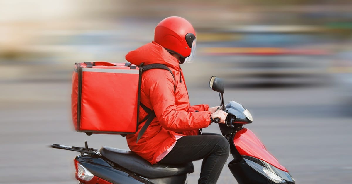 Kuvassa on ruokalähetti joka kulkee skootterilla päällään punainen takki ja punainen kypärä.