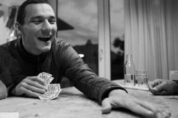 Kuva on väritön harmaa ja siinä esiintyy mies, joka istuu pöydässä ja hänellä on pelikortit kädessään.