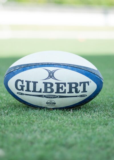 Kuvassa on sinivalkoinen rugby pallo, jossa lukee "girlbert" ja pallo on vihreällä nurmikolla auringon paisteessa.