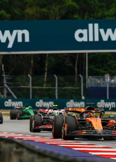 Kuvassa näkyy Formula 1 autoja kilpailemassa asfalttiradalla, jossa on punavalkoiset reunukset. Kuvan yläosassa on sinertävällä pohjalla mainoskyltti jossa lukee valkoisella Allwyn.