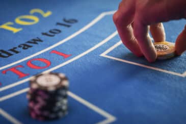 Kuvassa näkyy sinisellä kankaalla oleva Baccarat pöytä, jossa käsi asettaa pelimerkkejä.